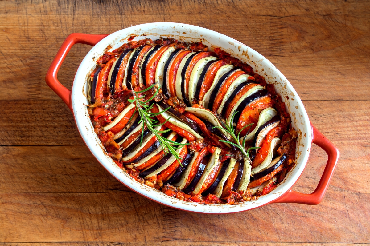 Collage of four dishes including a layered ratatouille in an oval casserole dish, crispy chicken cutlet with lemon slices on a black plate, a pot of creamy pink pasta and a plate of white spaghetti noodles garnished with basil leaves.